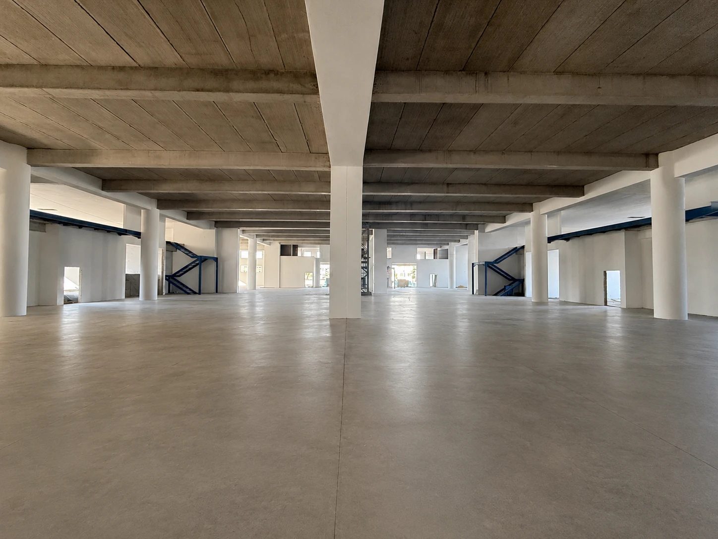 Rez-de-chaussée en chantier : grand plateau béton, poteaux blancs, escaliers métalliques bleus vers la mezzanine